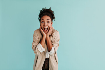 Obraz na płótnie Canvas Amazed black lady in trench coat looking at camera with smile. Front view of shocked african american woman posing on turquoise background.