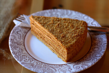 Delicious big triangular layered crumbly soft fragrant piece of honey cake on white plate with brown dots on wooden table, fork wrapped in napkin, side light from window, cozy cafe