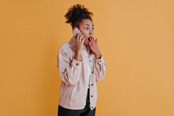 Shocked black lady talking on smartphone. Studio shot of surprised african american girl isolated on orange background.