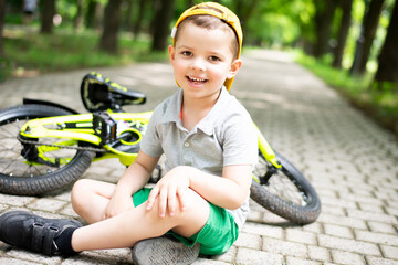 A small smiling five-year-old boy in a yellow cap sits on the path with his bright lemon yellow...