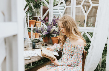 beautiful teen girl in a dress is reading a book from the list of school literature for the summer in a white veranda with flowers, a concept of summer vacation and simple living
