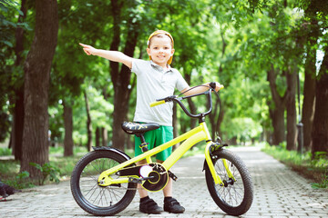 A small smiling five-year-old boy in a yellow cap stands on the path with his bright lemon yellow bicycle and points to the side.