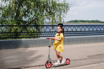 a charming girl rides a scooter along the river bank with a wrought-iron fence on a sunny summer day. 