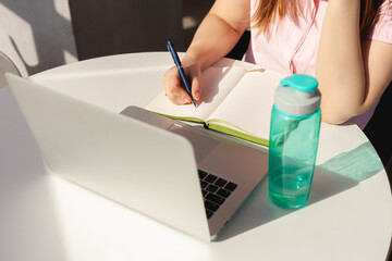 A girl writes with a ballpoint pen in a diary at her desk in front of a gray laptop in a bright...