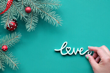 Christmas background on turquoise, green textile with wooden text Love. Top view on fir twigs decorated with red glass baubles, traditional Xmas toys.