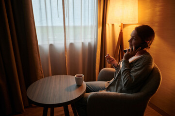Morning. Young woman in the living room speaks on the phone. There is a cup of coffee on the table in front of her. Beautiful interior. The windows have white and yellow curtains.