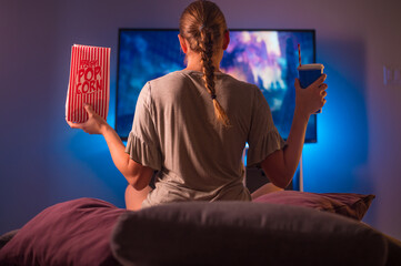 Relaxing evening. Watching your favorite TV shows. A young woman with blond hair sits on a couch in the bedroom and watches TV. Rest, relaxation. The woman is holding a pack of popcorn.