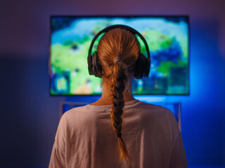 A young woman is engaged in gaming. Plays video games. The woman is wearing headphones. In front of her is a computer. Close-up. Shooting from the back.