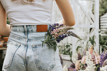 Summer bouquet of lupins in the pocket of jeans of a teenage girl in jeans and a white T-shirt stands on a white veranda with flowers, a concept of summer vacation and a simple living