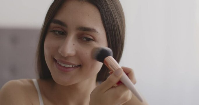 Young attractive middle-eastern lady applying rouge on her face with make-up brush, finishing everyday makeup at home