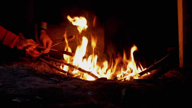 Hungry Tourists Grilling Sausages on Wooden Skewers, Sits by Night Bonfire. Bright flame with smoke from a fire in forest. Dry branches burn in open fire. Rest on nature. Tourism summer. Camping. Zoom