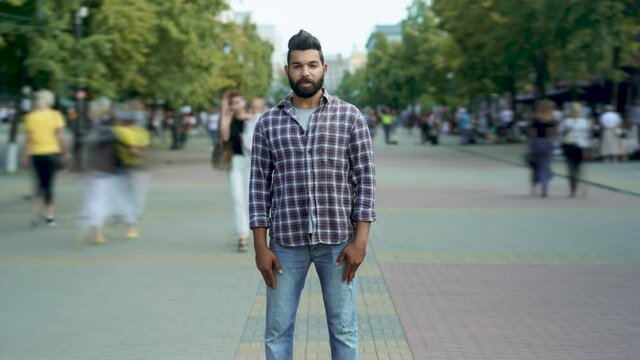 Zoom-in time lapse portrait of attractive Middle Eastern man standing alone among people passing by and looking at camera with serious face. Youth and society concept.