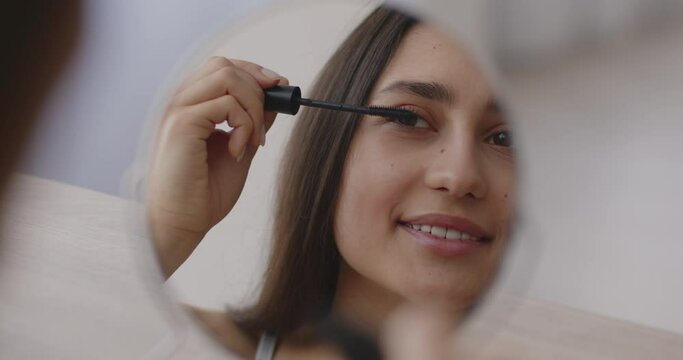 Light everyday makeup. Young middle eastern woman applying mascara on eyelashes and smiling, mirror reflection portrait