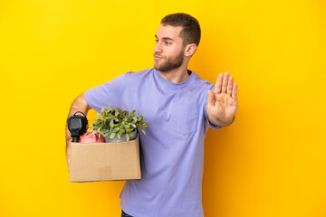 Young caucasian making a move while picking up a box full of things isolated on yellow background...