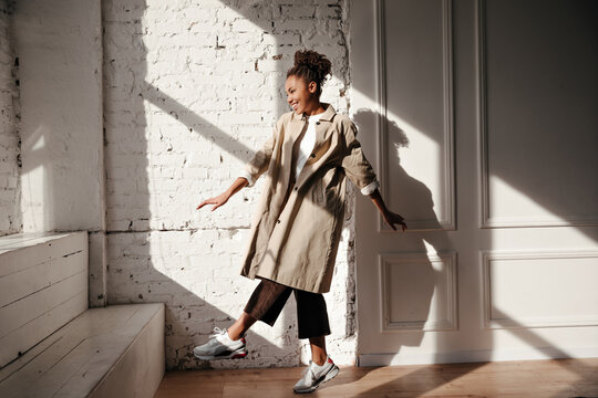 Full length view of pleasant black woman in trench coat. Laughing african american girl dancing under sunlight.