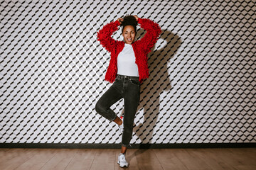 Full length view of blithesome black woman standing on one leg. Studio shot of african american girl in red jacket posing on textured background.