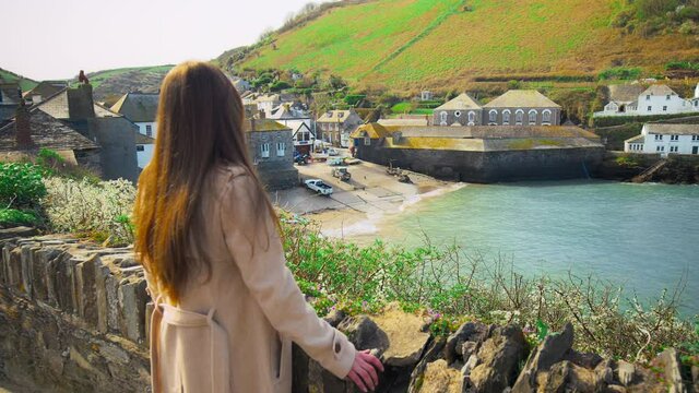 A girl looking at Port Isaac, fishing village, Atlantic coast Cornwall England. A young woman watching at Porthysek, rocky scenic views with blue waters, green lands and old English houses in 4K.