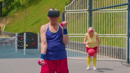 Obraz premium Active senior man grandfather in VR headset doing sport training fitness stretching aerobics cardio exercising with dumbbells on playground. Healthy grandmother playing ball basketball on background