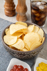 Potato chips in a bowl, pepper, salt, mustard, ketchup and a glass of soda