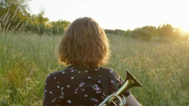 the view from the back, a young creative woman with a trumpet behind her back is walking through a field towards the sun in a summer dress. The image of a freedom-loving, unique person open to new