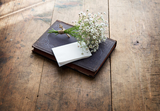 Vintage book and a small bouquet of baby's breath flowers and a blank sheet of folded white memo paper, on a well used old desk or wooden surface.