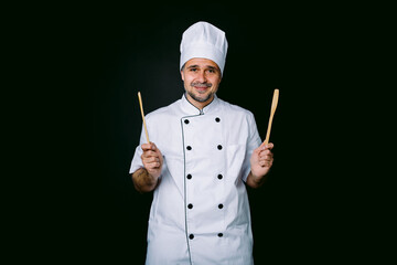 Chef cook wearing cooking jacket and hat, holding wooden spoon and fork in his hands, on black background