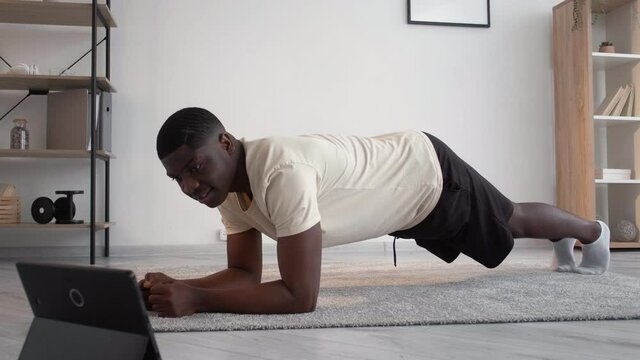 Online training. Sportive black man. Home fitness. Healthy lifestyle. Smiling african guy black shorts white t-shirt doing plank workout looking portable computer light room interior motion view.