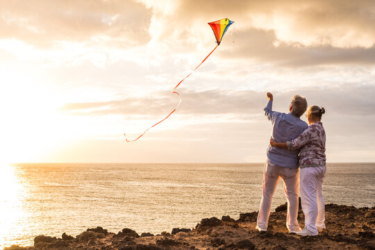 close up and portrait of two old and mature people playing and enjoying with a flaying kite at the beach with the sea at the background with sunset - active seniors having fun.
