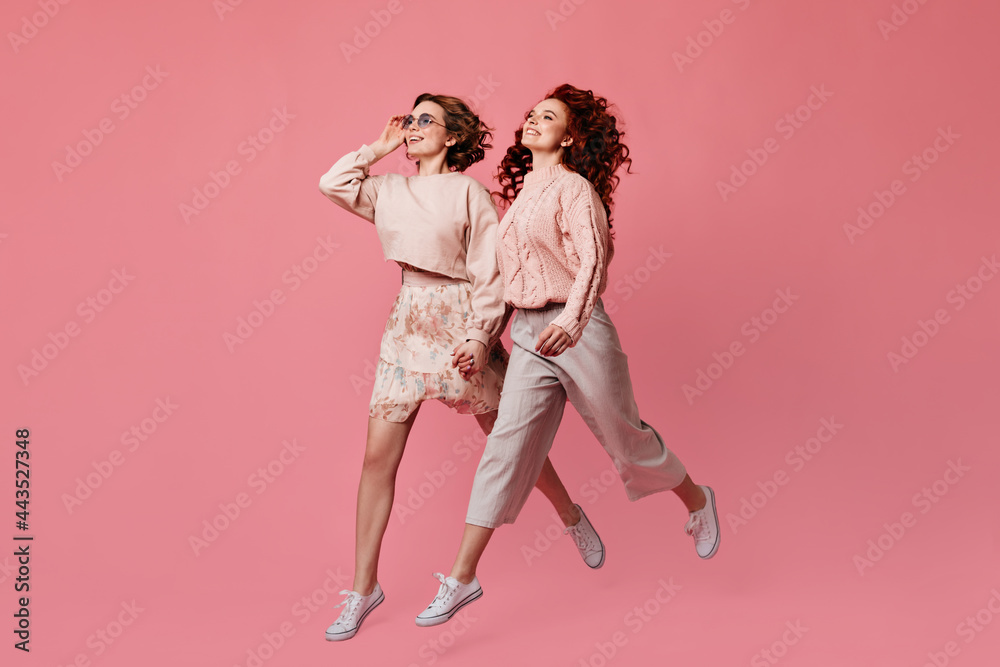 Sticker Two laughing girls holding hands. Studio shot of female friends running on pink background.