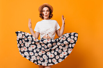 Amazed european woman in trendy skirt posing on yellow background. Studio shot of emotional fashionable girl in straw hat dancing with surprised face expression.