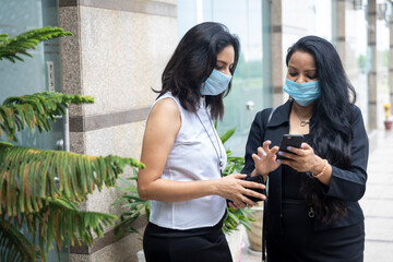Two Indian woman wearing Covid-19 protection mask look into their phone in an urban corporate setting.