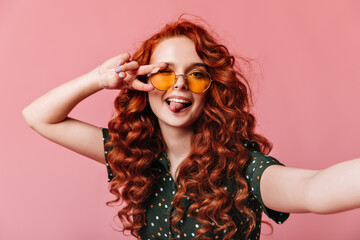Positive ginger girl taking selfie with peace sign. Pretty red-haired lady making funny faces on pink background.