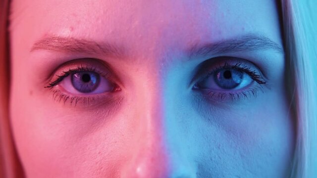 Beautiful young woman opens and closes her eyes in color lighting close-up. Large blue female eyes in neon light. Slow motion, HD.