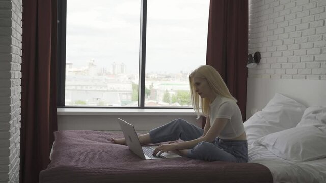 Young woman works at a laptop in a cozy bright bedroom. The blonde woman is typing on the keyboard against the background of a large window. Remote work concept. 4K UHD
