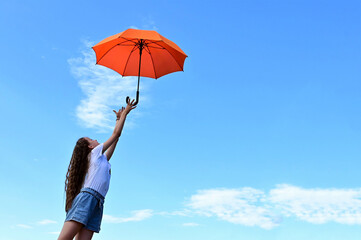 beautiful young girl white T-shirt and denim shorts throws up an orange umbrella. A flying umbrella against the blue summer sky. A missed chance. Copy space