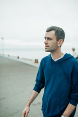 middle-aged man walks on the sea pier. portrait of young man with dark but already partially gray hair in blue hoodie near the sea. urban style content. selective focus