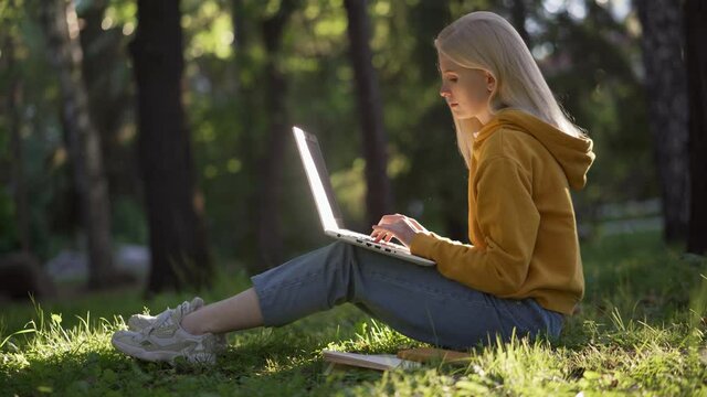 A young blonde woman in casual clothes with a laptop sits on the green grass. The sun illuminates the laptop. Books are lying nearby. Study in nature. 4K UHD