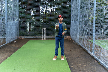 Active young boy bouncing cricket ball. South Asian look child playing with red cricket ball on cricket pitch under nets.
