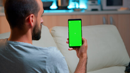 Relaxed man looking at smartphone with mock up green screen chroma key display while relaxing on sofa. Caucasian male holding in horizontal mode phone with isolated display late at night in kitchen