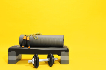 Step platform, dumbbell, mat and jump rope on yellow background, space for text. Sports equipment