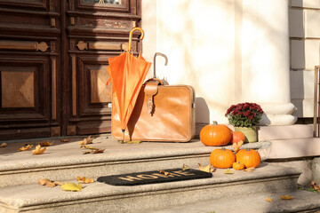 Doormat with umbrella, suitcase and pumpkins near entrance of house on autumn day