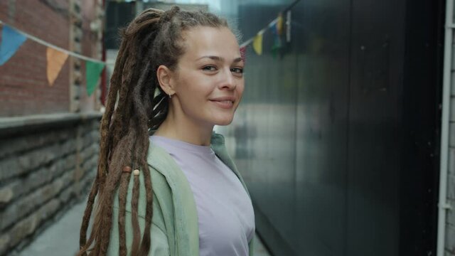 Portrait of good-looking young lady with dreadlocks turning to camera and smiling outside in city. Extraordinary people and subculture concept.