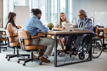 Selective focus of smiling disabled businessman and colleagues in office. Side View Of A Disabled Businessman Sitting On Wheelchair Using Laptop Working In Office. Multicultural businesspeople