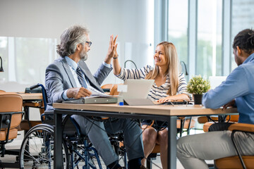 Selective focus of smiling disabled businessman and colleagues in office. Side View Of A Disabled Businessman Sitting On Wheelchair Using Laptop Working In Office. Multicultural businesspeople