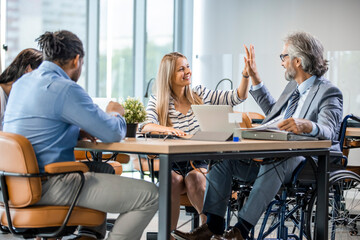 A man in a wheelchair is working in a bright office. His colleagues are passionate about work. Disabled Male Manager Sitting With His Colleagues At Workplace. Group of business people in a meeting