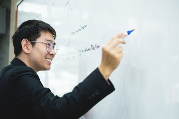 An Asian male teacher in a classroom near the whiteboard is writing and teaching a lesson with a special lecture in a class column. Math teacher explains arithmetic sums to university students
