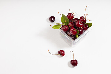 Fresh juicy red cherries in plastic container with green leaves, tails in sunlight as summer healthy food on white wood table, top view, border, copy space.
