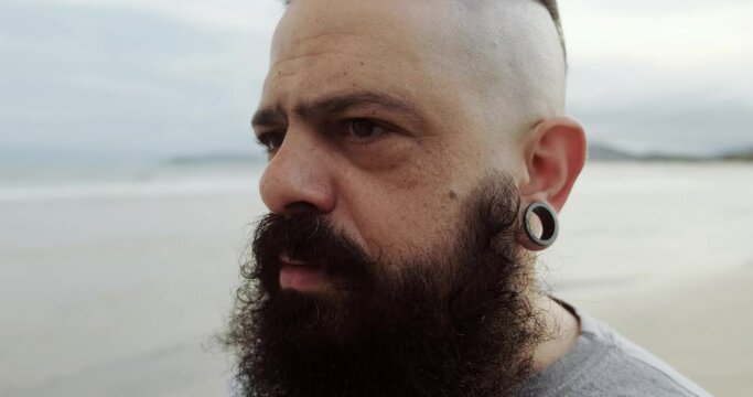 Serious latin man with big beard on a beach looking at horizon. 4K.