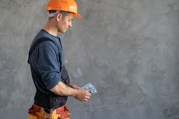 A builder in an orange construction helmet is counting dollar bills while standing near the wall....