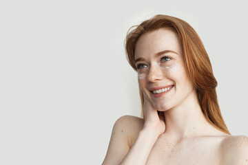 Obraz premium Woman with freckles and red hair is posing on a white studio wall applying on a her face anti aging cream